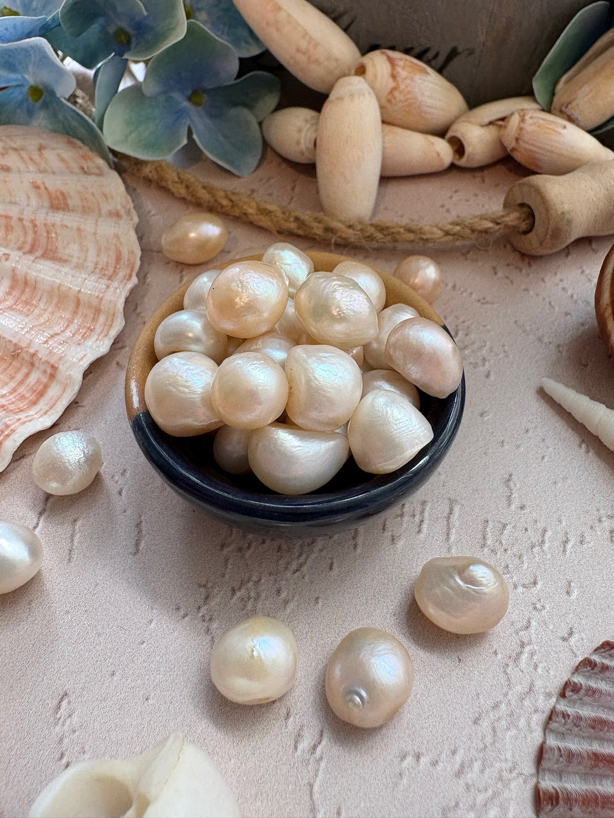 Freshly cultivated white pearls in a small dark bowl, surrounded by seashells and decorative elements on a textured beige surface.