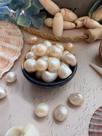 Freshly cultivated white pearls in a small dark bowl, surrounded by seashells and decorative elements on a textured beige surface.